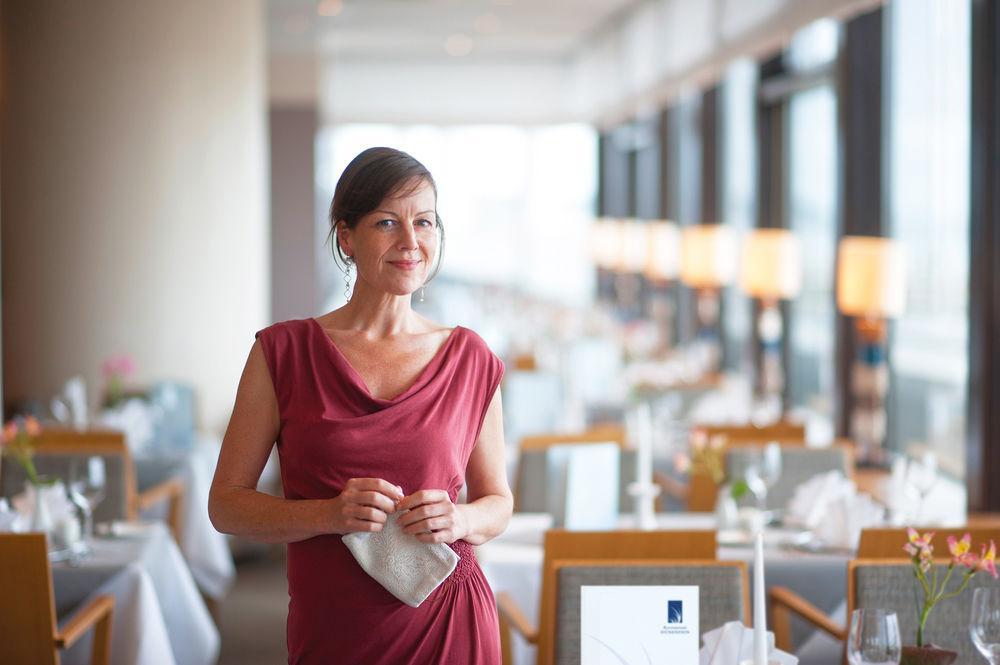 Hotel Neptun Rostock Exterior foto The photo shows a woman standing in a stylish restaurant. She is wearing a red dress and holding a small clutch. The background features a well-lit dining area with tables set for meals, including white tablecloths, elegant dinnerware, and decorative